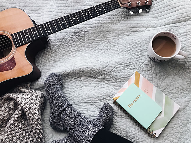 Person sitting on a bed with coffee, book and guitar